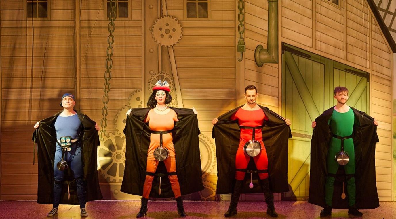 Four performers stand on a stage in a row facing forward. Each one is wearing a colourful bodysuit and long black coats that they are holding open to reveal their costumes. Attached to the front of each bodysuit is a kitchen pot or pan, positioned at waist level. One of the performers also wears a decorative hat.