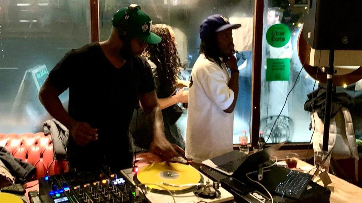 Rapper Talib Kweli performs a DJ set at The Social in Stokes Croft in Bristol. He is wearing a black T-shirt and black cap and is spinning records on his decks, with other technology visible. Two other people are stood behind him looking out into the crowd