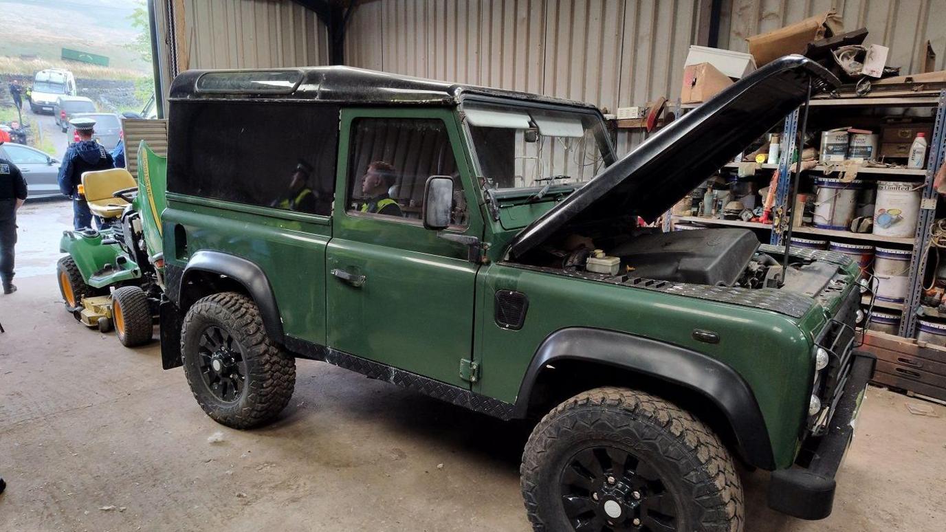 An green Land Rover with an open bonnet stands in a shed, in front of a small green vehicle