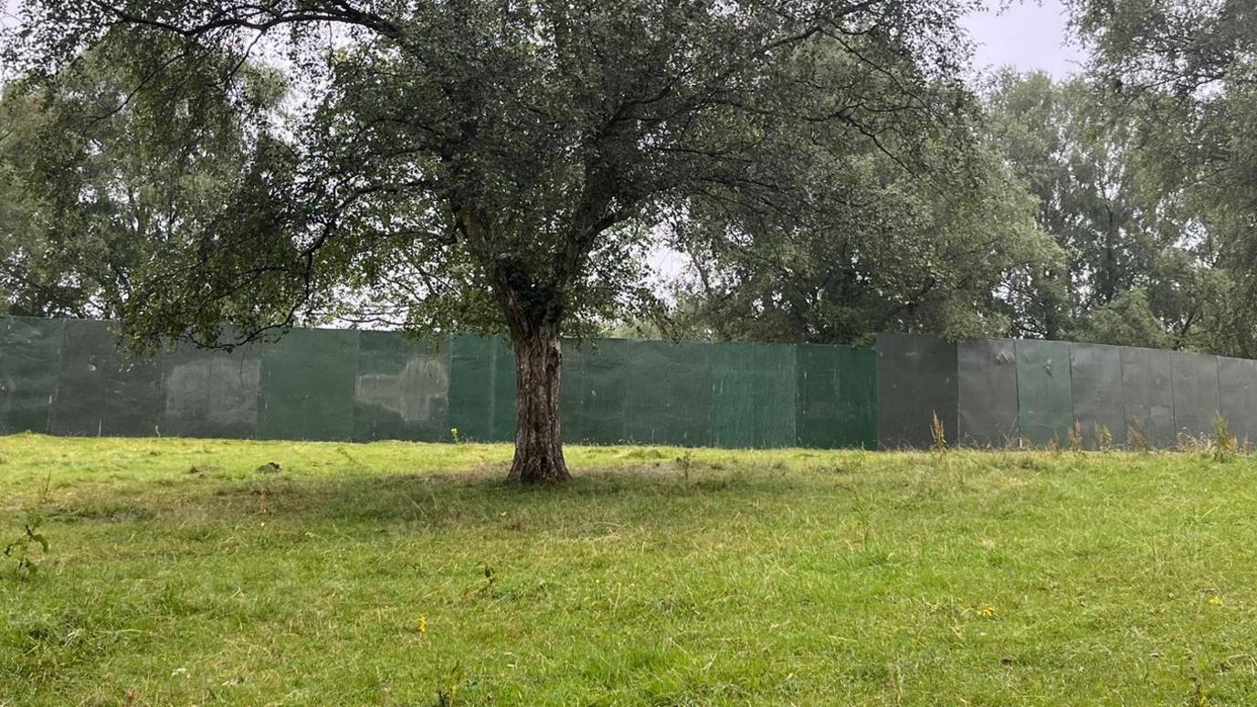 A large green fence erected across a green field with tree seen in the foreground.