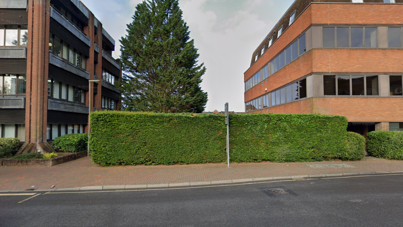 The entrance to the footpath that is the start of Bissengen Way in Knoll Road, Camberley. A box hedge is in the foreground and red brick office buildings in the background.