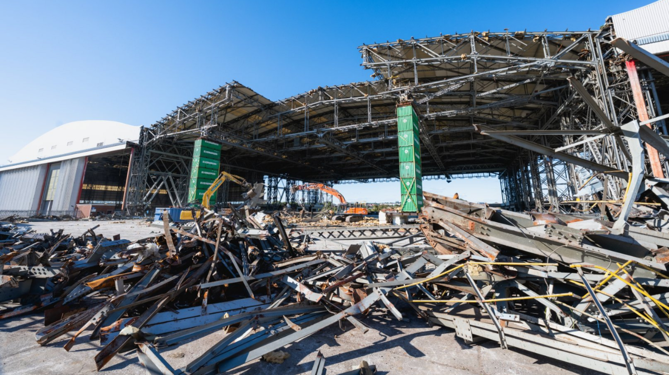 Ground view showing the hangar mid demolition. The structure is partly supported by green Maersk branded shipping containers stacked on top of each other and piles of metal debris in the foreground.