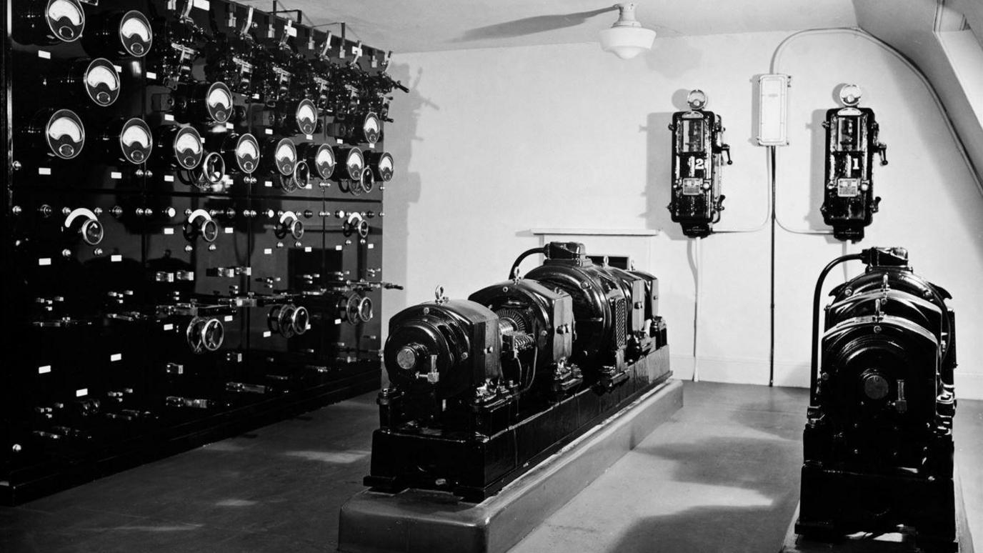 A black and white picture of a control room. One wall is completely filled with dials and switches, and there are two rows of old equipment sitting in the middle of the floor.