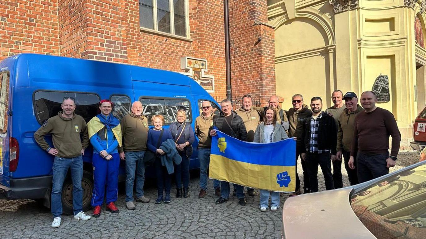 A group of people stood in front of a blue van with two people holding a blue and yellow flag. 