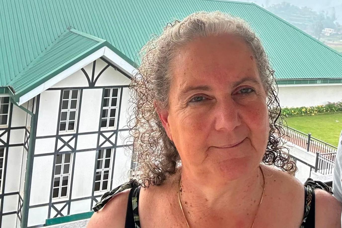 A woman in her 60s with grey curly hair pulled back from her face stands in front and slightly above of a large white building with black window frames and a green roof. She is wearing and has tie straps on her shoulders. In the distance a misty hill side is in view.