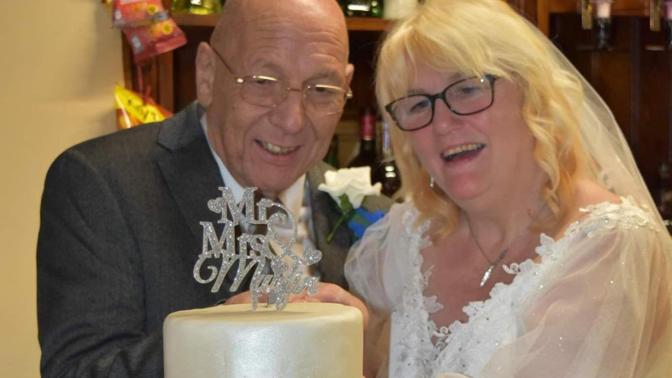 Steven and Julie Murfin cutting the cake on their wedding day. He is wearing a dark grey suit, white shirt and a tie, with a white rose pinned to his lapel. She is wearing a wedding dress and veil