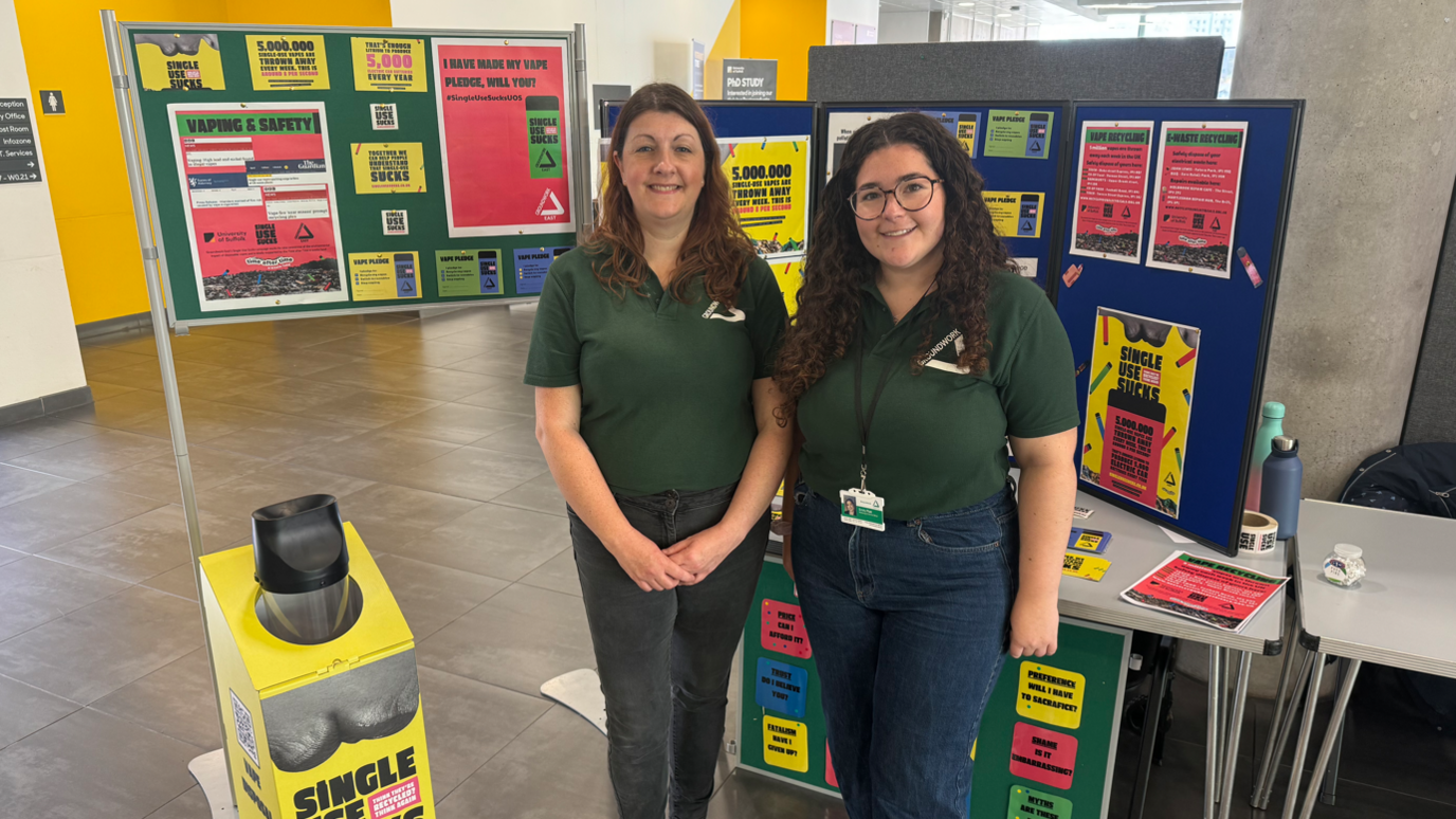 Two women wearing dark green polo shirts standing in front of boards on which there are posters about a charity's Single Use Sucks campaign against disposable vapes