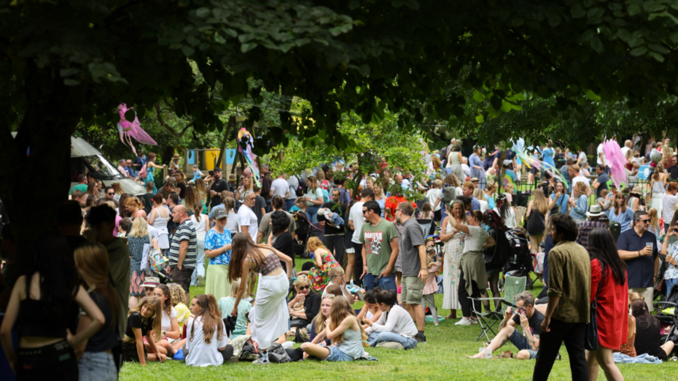 A park filled with people standing and sitting on the grass.