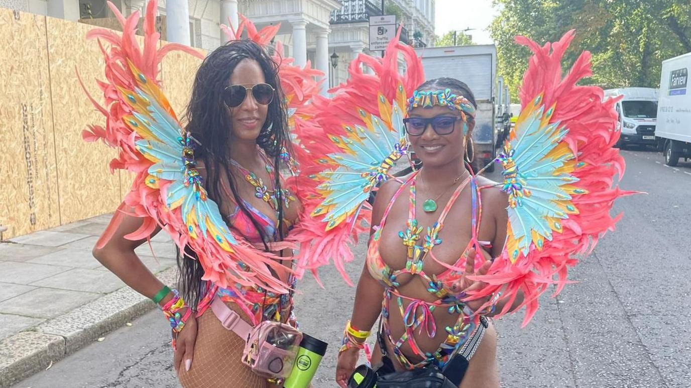 Laila and Candice at the Notting Hill Carnival are dressed in vibrant, feathered costumes with large, colourful wing-like accessories on their backs. Both wear bikini-style outfits adorned with pink, blue, and yellow feathers. One has long braids and sunglasses; the other wears glasses with a matching headpiece. 