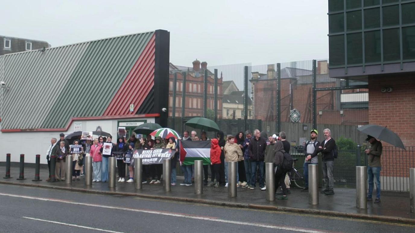 A group of about 30 pro-Palestinian protesters gathered outside Musgrave Street PSNI station in Belfast city centre. Some are holding banners and umbrellas. A large Palestinian flag is being held up in the middle of the group.