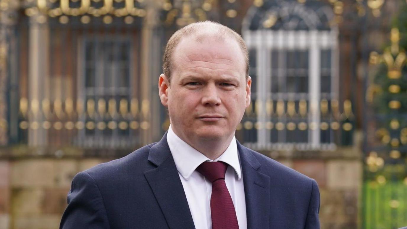 Headshot of Minister of Communities, Gordon Lyons, wearing a navy suit, white shirt and burgundy tie. 