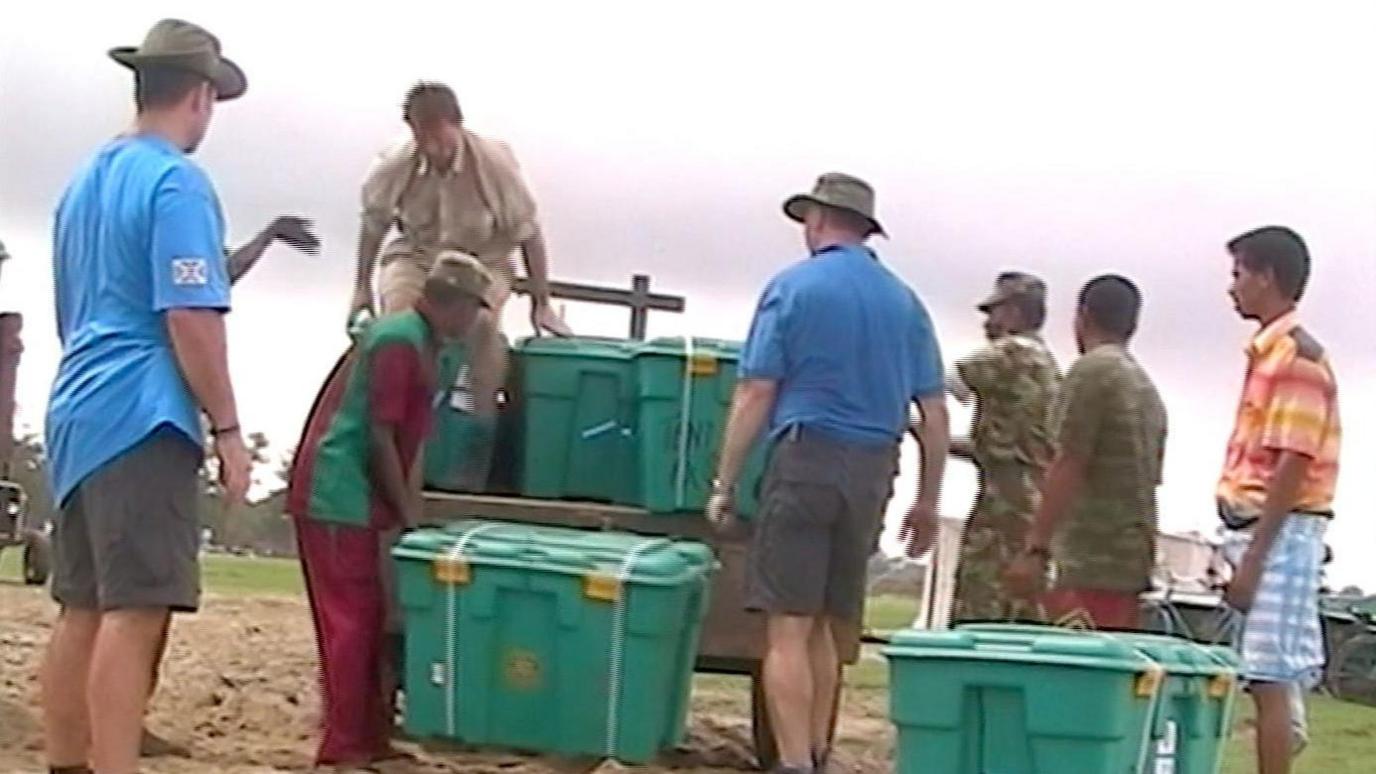 A group of men pull large green plastic boxes off a cart.