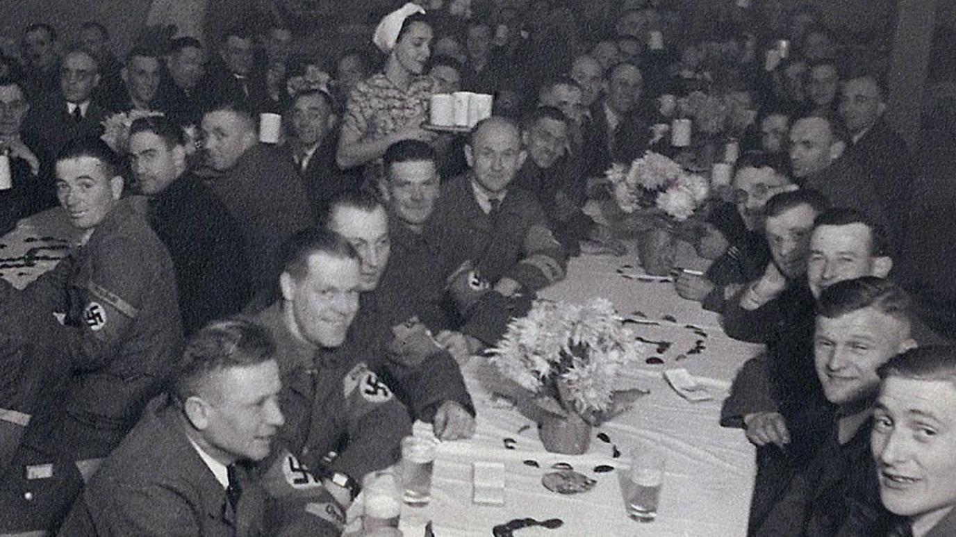 Uniformed German soliders with swastika arm bands sit around long tables drinking from glasses and mugs