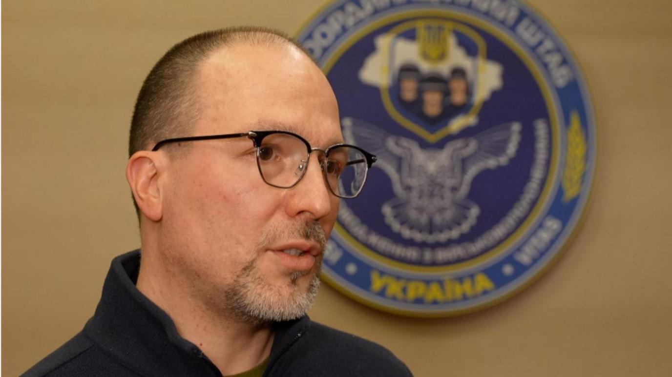 A man wearing glasses in front of the emblem of the Ukrainian military.