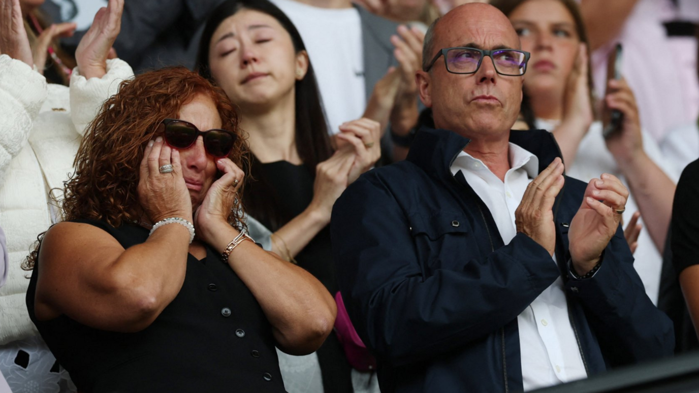 Jota's mother and father are wearing dark clothing and are both emotional during  the tribute. Isabel has her hands on her faces and is crying behind sunglasses. Joaquim has a white short on under a jacket and glasses. He is clapping.