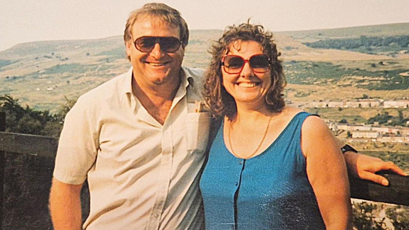 A photo of Jayne as a younger woman with her late husband. They are posing together at a viewpoint. Jayne has long curly hair and a blue dress and her husband is wearing a beige short-sleeved shirt.