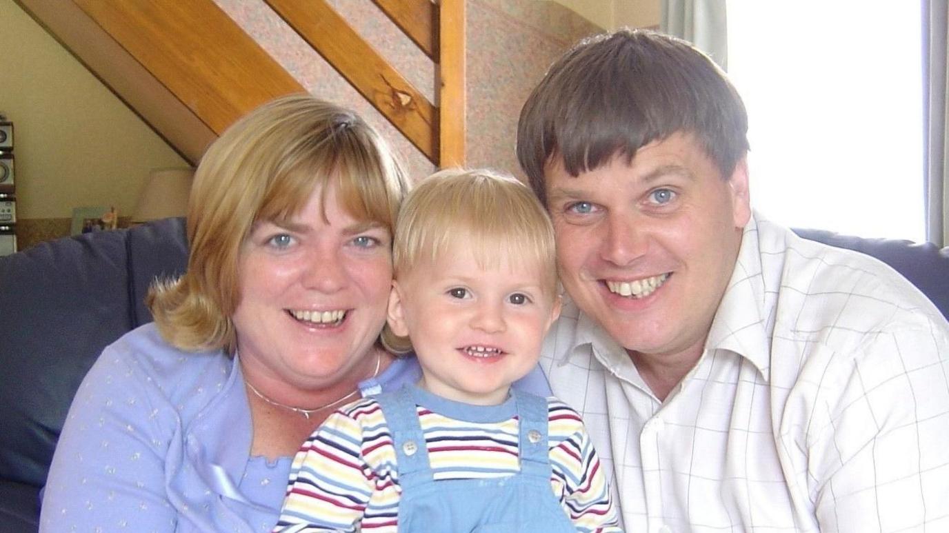 James Eyn eistedd gyda'i rieni Ruth ac Andrew Evans. Mae James yn gwisgo cris hir streipiog. Mae'n gwisgo dungarees glas ac mae ganddo wallt melyn. Mae Ruth yn gwisgo top glas. Mae ganddi wallt melyn. Mae hi'n gwisgo mwclis. Mae Ruth yn gwenu ac mae ganddi lygaid glas. Mae Andrew yn gwisgo crys gwyn gyda sgwariau llwyd. Mae'n gwenu ac mae ganddo wallt brown a llygaid glas.