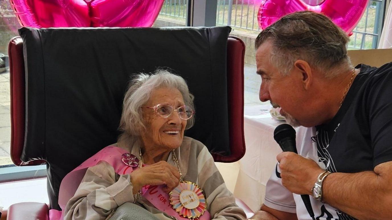 Ada Cook is on the left sat on a black and red chair. She has shoulder length grey hair and is wearing a pink birthday sash and a badge. On the right is a man wearing a white and black T-shirt holding a black microphone. 