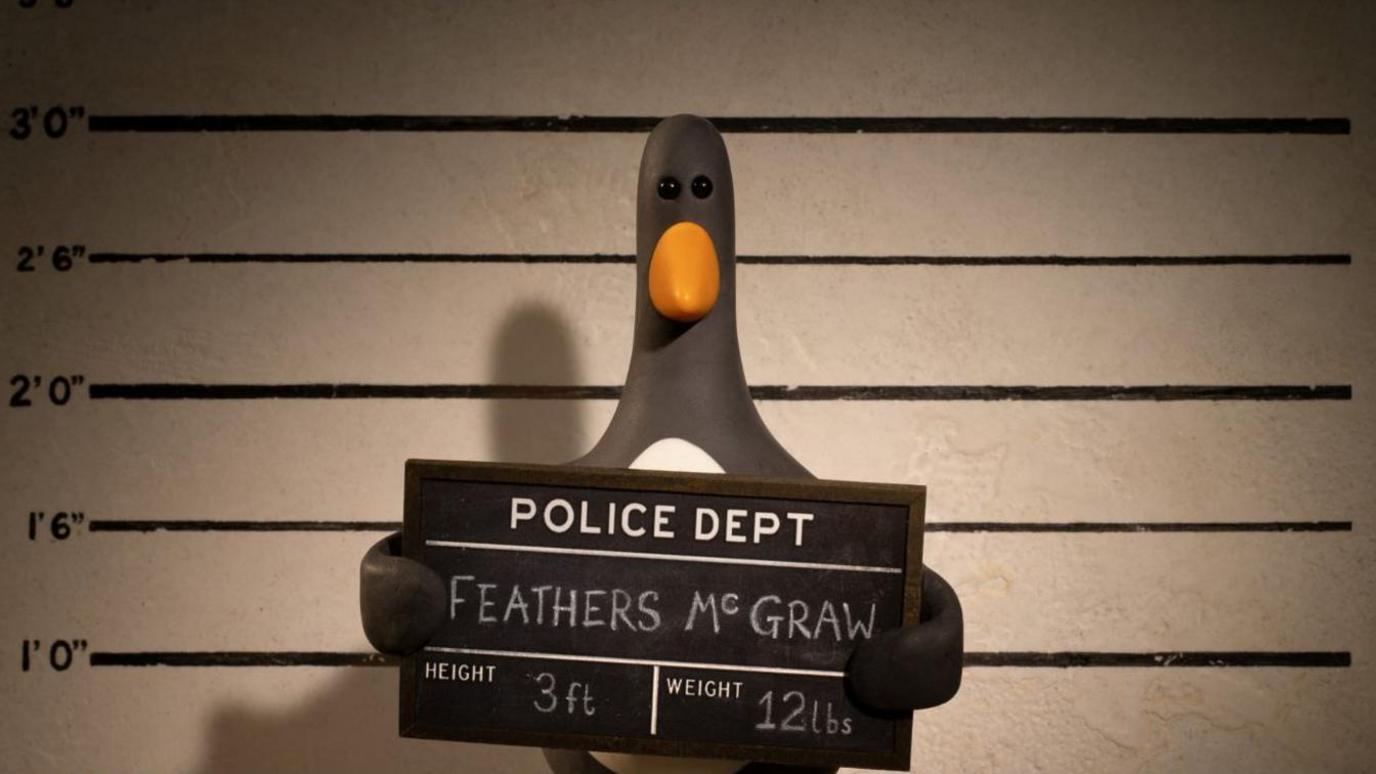 A plasticine model of a penguin poses for a police mug shot. He holds a board reading Police Dept: Feathers Mc Graw; Height: 3ft; Weight: 12lbs.