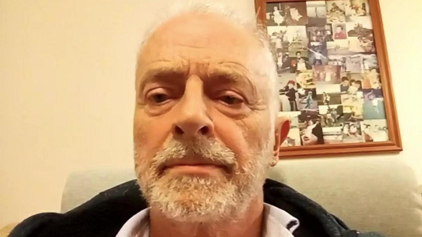 Chris Mullen with short white hair, and a white moustache and beard, sitting in a chair in a living room. There's a frame of family photographs behind him on a wall.