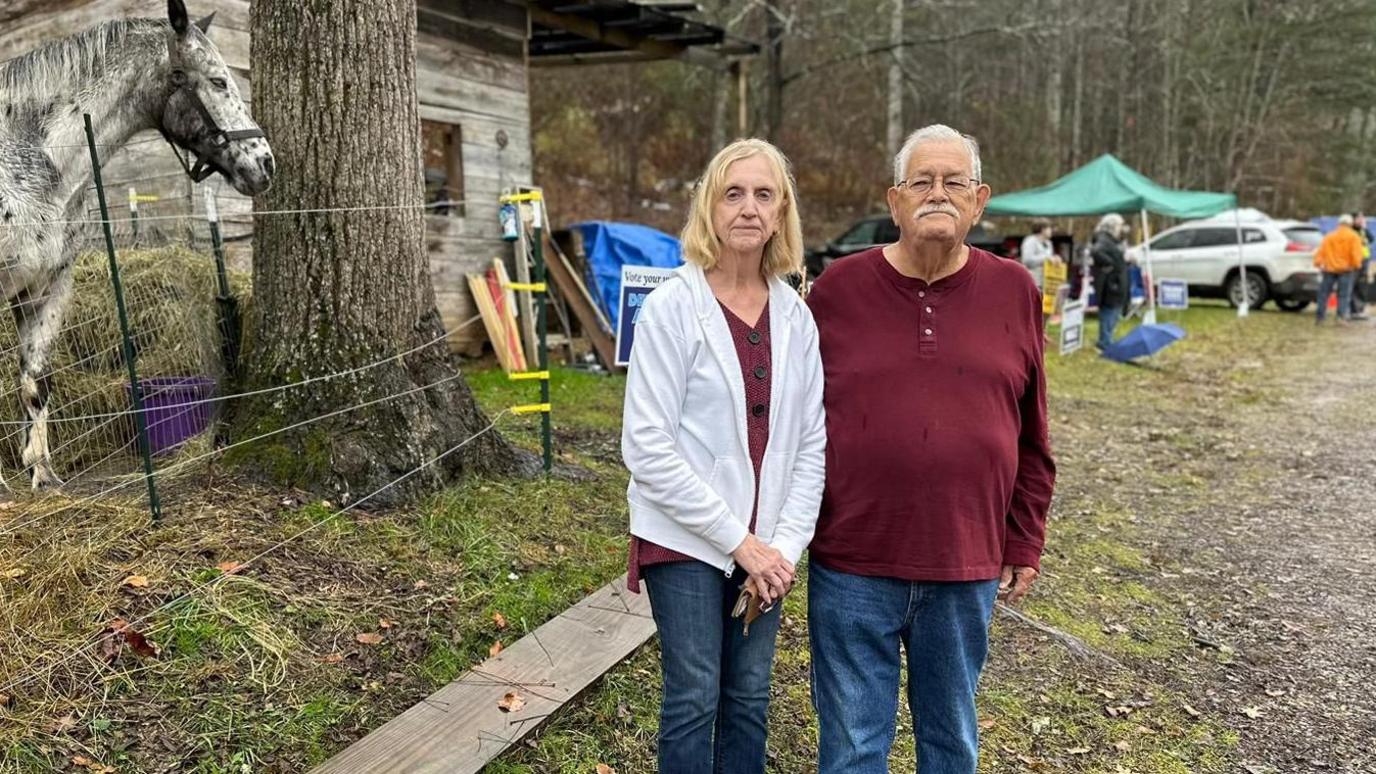 Man and woman in red shirts with jeans on gravel road next to horse, weathered building green canopy tent and cars.