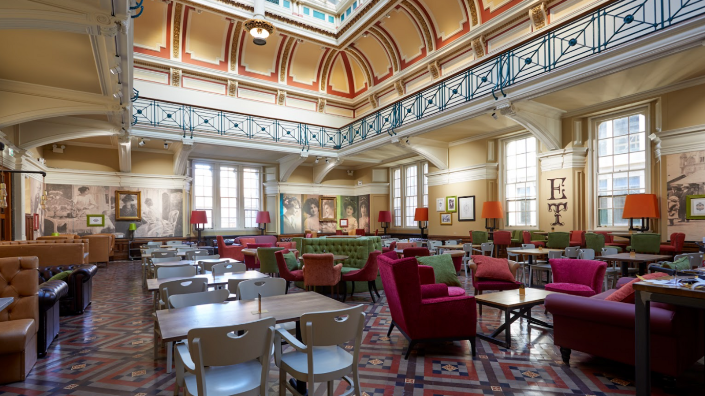 Chairs and tables fill a bright, spacious room, the walls are cream, except for the back wall which has portraits and photographs covering it