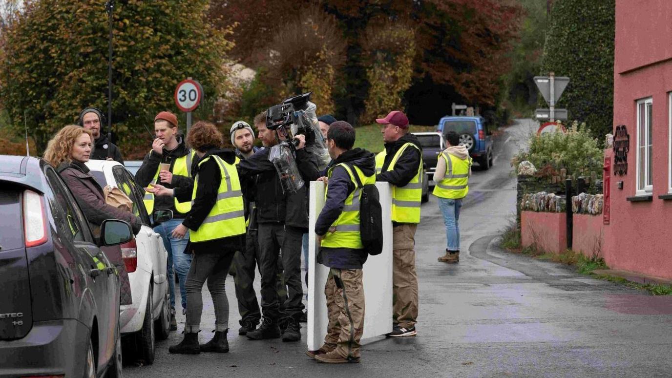 A film crew in high-vis vests is gathering around a woman stood next to a line of cars. The woman appears to be an actor. They are on a street in a country village, with a red house to the right. There are large trees in the background.
