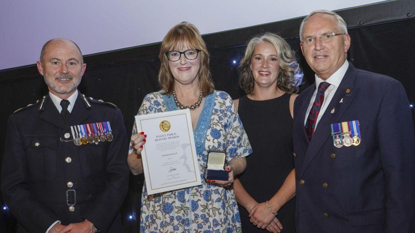 Gavin Stephens, Elizabeth Powell, Hannah Conroy and Chris Nelson are pictured standing together. All are smiling and Elizabeth is holding a certificate and a medal. She is wearing a blue and white floral-patterned dress. The two men are wearing suits with medals attached to them. PC Conroy is wearing a black dress.