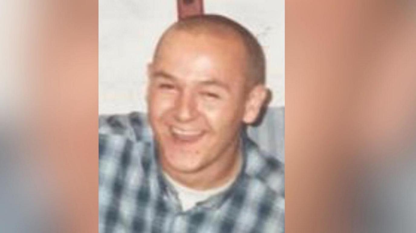 Vincent Lawrence smiles as he is photographed. He is wearing a blue-and-white checked shirt, buttoned up over a white t-shirt.