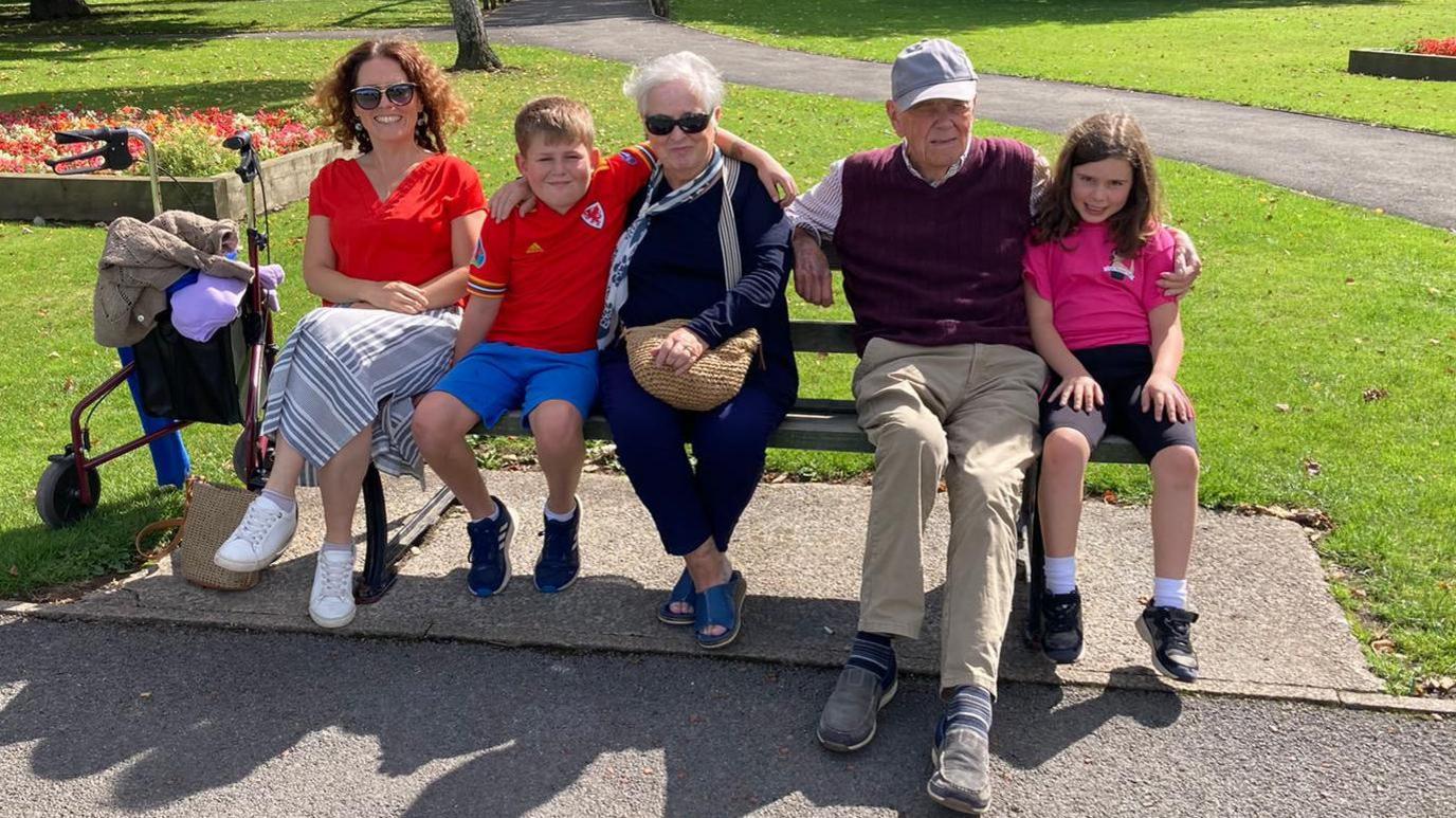 A family sat on a bench in a park, with a walker parked next to them. From left to right: A woman with brown hair and sunglasses, in a red top and grey striped skirt, next to a young boy in a Wales football shirt and blue shorts, who has his arms around a woman with short grey hair dressed all in dark blue with a white scarf and brown handbag. Next to her is an older man in a grey cap, purple sweater vest and beige trousers. He has his arm around a young girl with brown hair in a pink top
