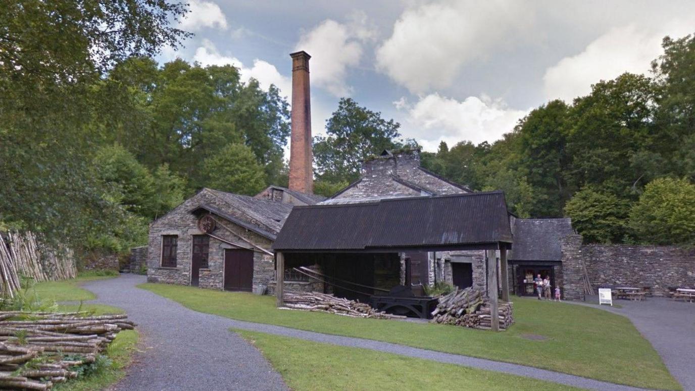 Mill building in greenery. It is made of grey brick. 