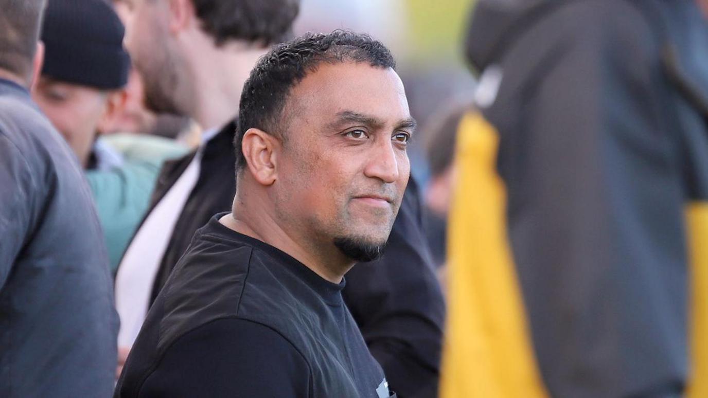 A side profile of George Akhtar, who is standing in a crowd of people. He has dark hair and is wearing a black jumper.