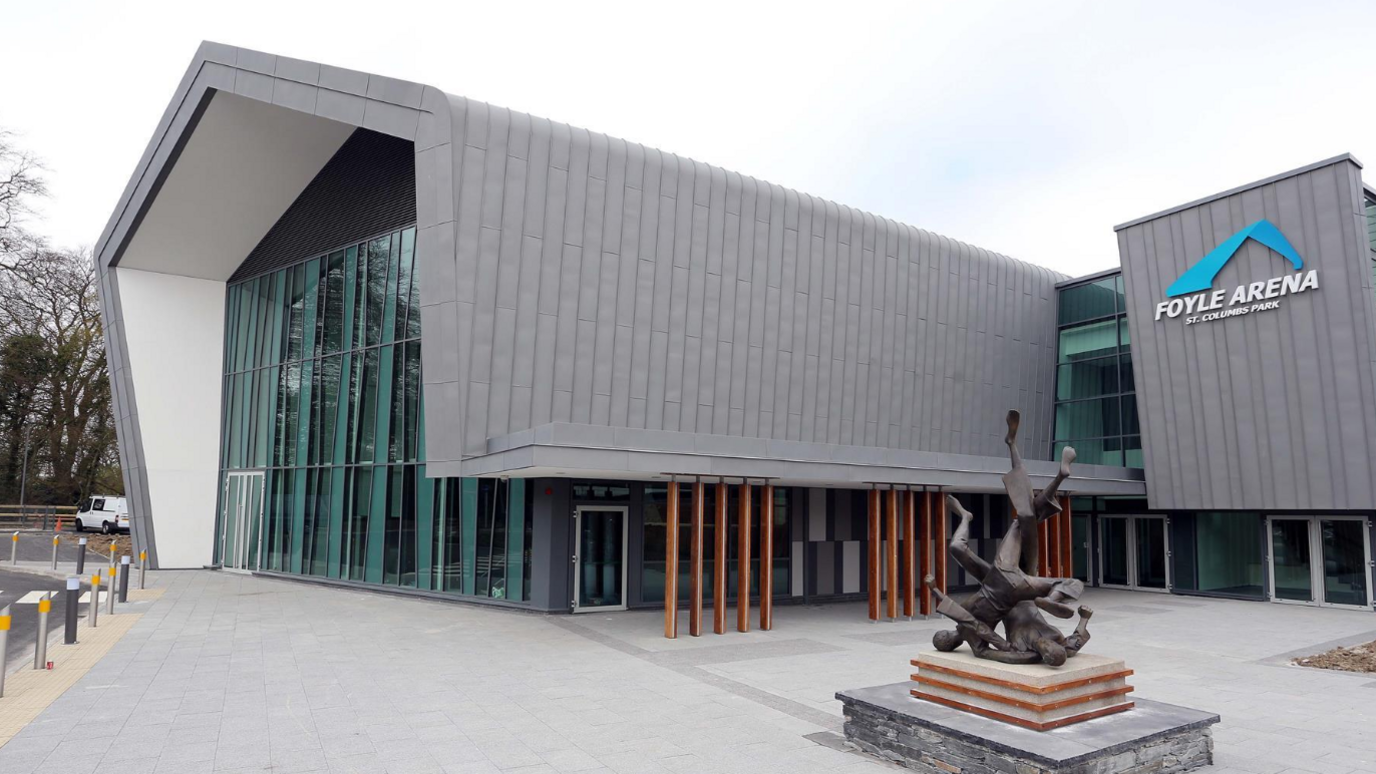 Foyle Arena is a large grey building with a statue near its entrance.
It has a brown columns and a large window display near it's front.