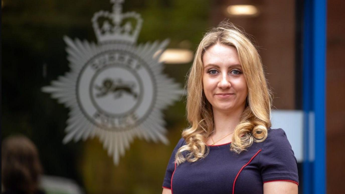 Ellie Vesey-Thompson, a young woman with long blonde hair and wearing a blue dress with red piping, stands in front of a glass door with the logo of Surrey Police on it.