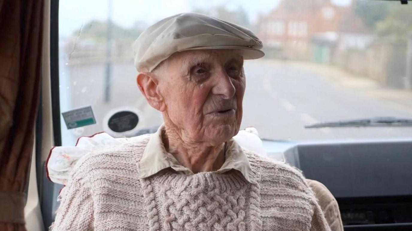 Bill wearing a cream cap and a beige jumper and shirt sat inside a van with the road behind him