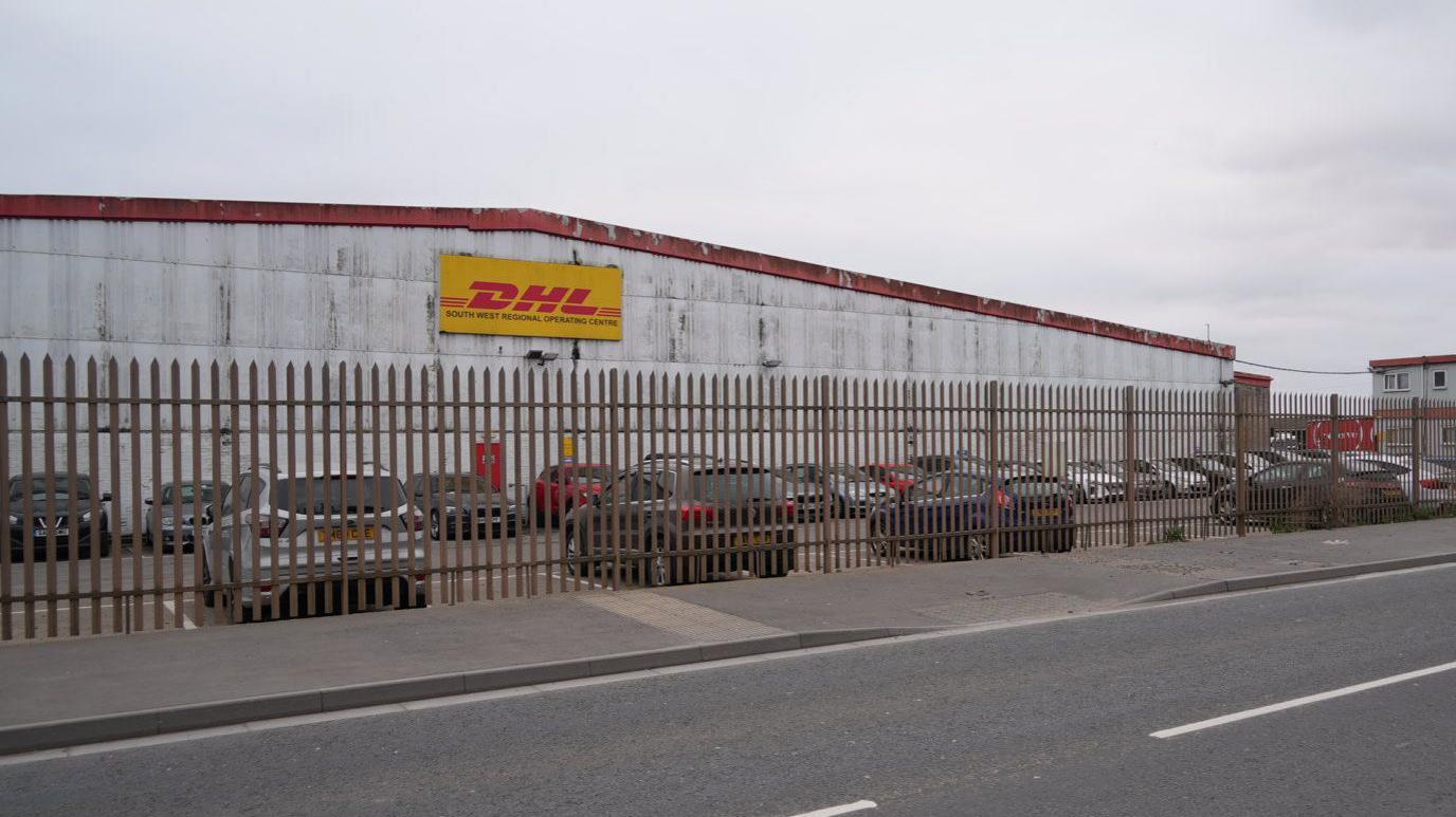 The DHL warehouse in Avonmouth, pictured from across a road. There is a tall metal fence running around the outside of a car park, and the structure itself is clad with white corrugate iron, heavily weathered. There is a DHL sign in yellow and red on the side of the building, and a faded red corrugated iron roof. 