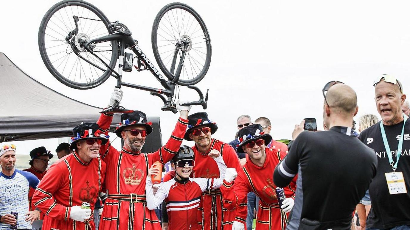 Four men in Beefeater costume holding aloft a bicycle, and a young cyclist with raised arms in front of them. A man in lycra cycling wear takes a photo of them on a mobile phone while other people look on