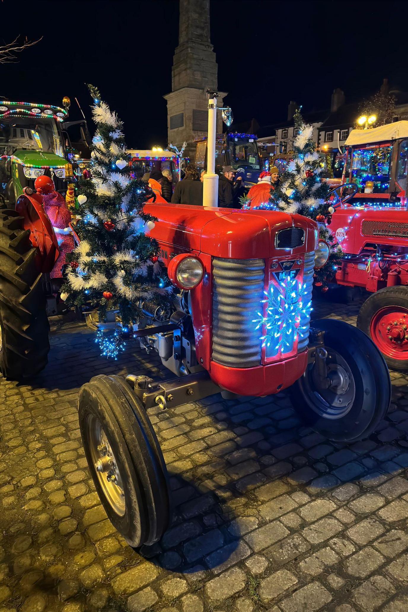 A vintage red tractor covered in tinsel, lights and Christmas trees in Ripon's market square at night 