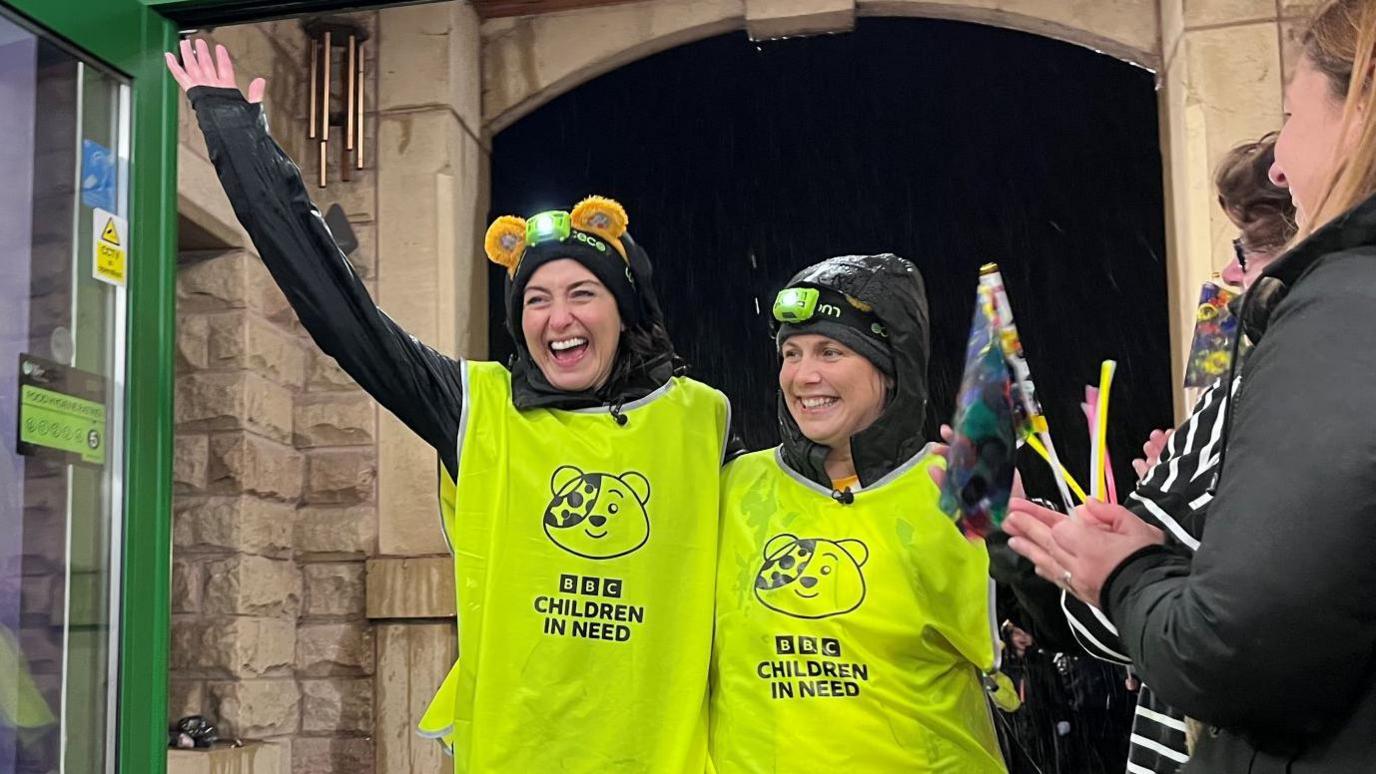 Two smiling woman stood closely next to each other in a doorway, one with their arm raised in the air. They are wearing high-vis vests which read 'BBC Children In Need'.