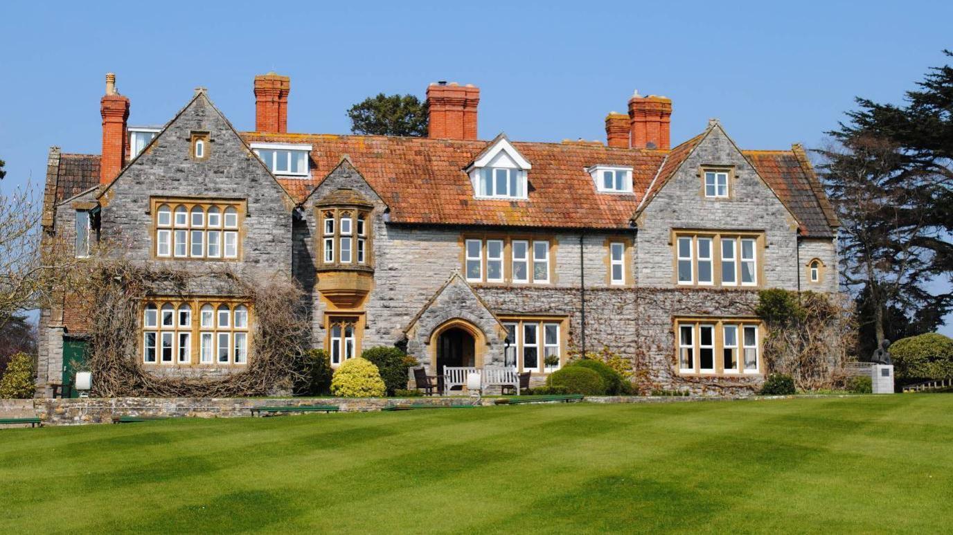 A large historic looking boarding school with grey pointed bricks, terracotta tiled roof and lots of bay windows. There is a bright green manicured lawn in the foreground, and various climbing plants on the side of the building.