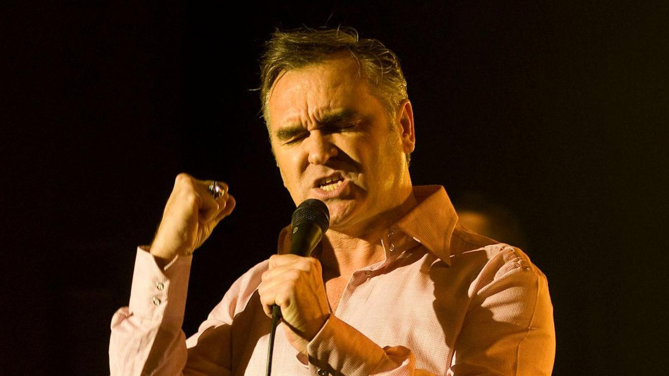 Morrissey on stage holding a microphone, clenching his first with his eyes closed as he sings against a blacked out backdrop.