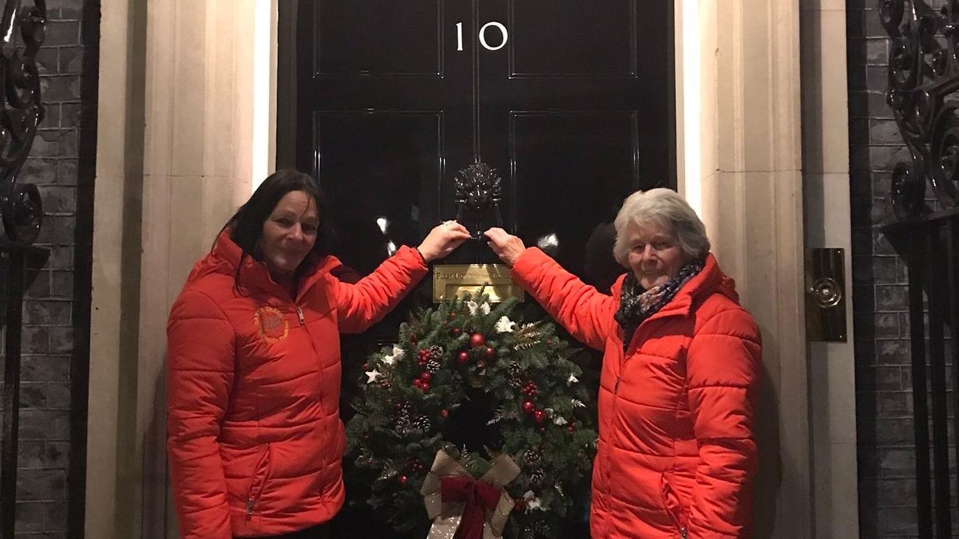 Two women in red coats knocking on Number 10 Downing Street with their champion wreath 