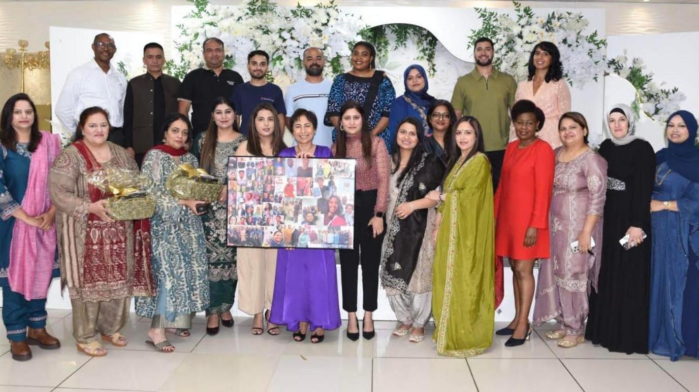 Image shows a group of people celebrating 10 years since Love in Care's launch. Aqila Choudhry is featured in the centre and is wearing a purple dress and is holding a photo frame. Other employees are gathered around her.