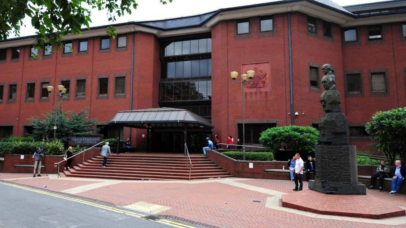 A large, three-storey red brick building with a canopy over the glass front entrance. People are milling about and there is a statue with four heads on it to the right hand side.