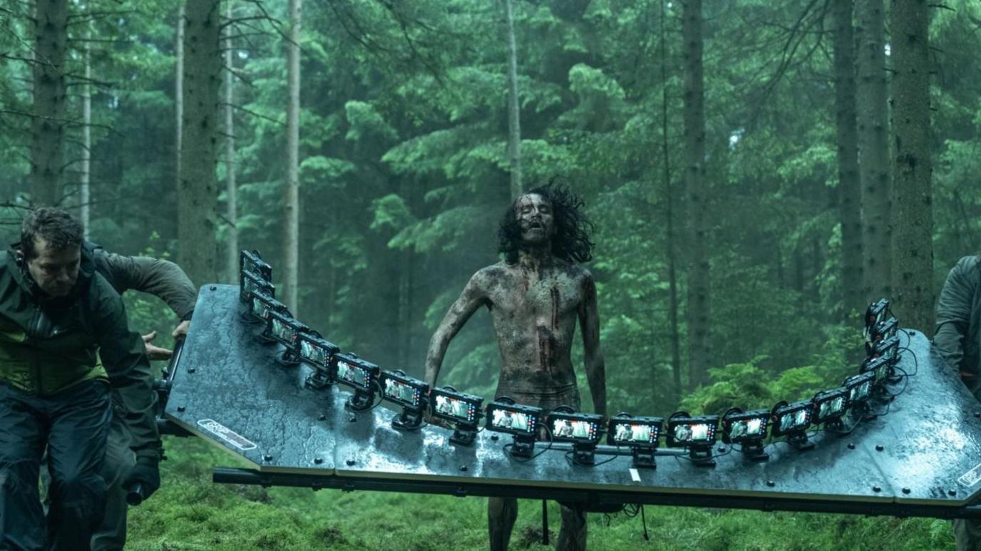 A man made up to look like a zombie runs through a forest with film crew members in front of him holding a sheet of lights