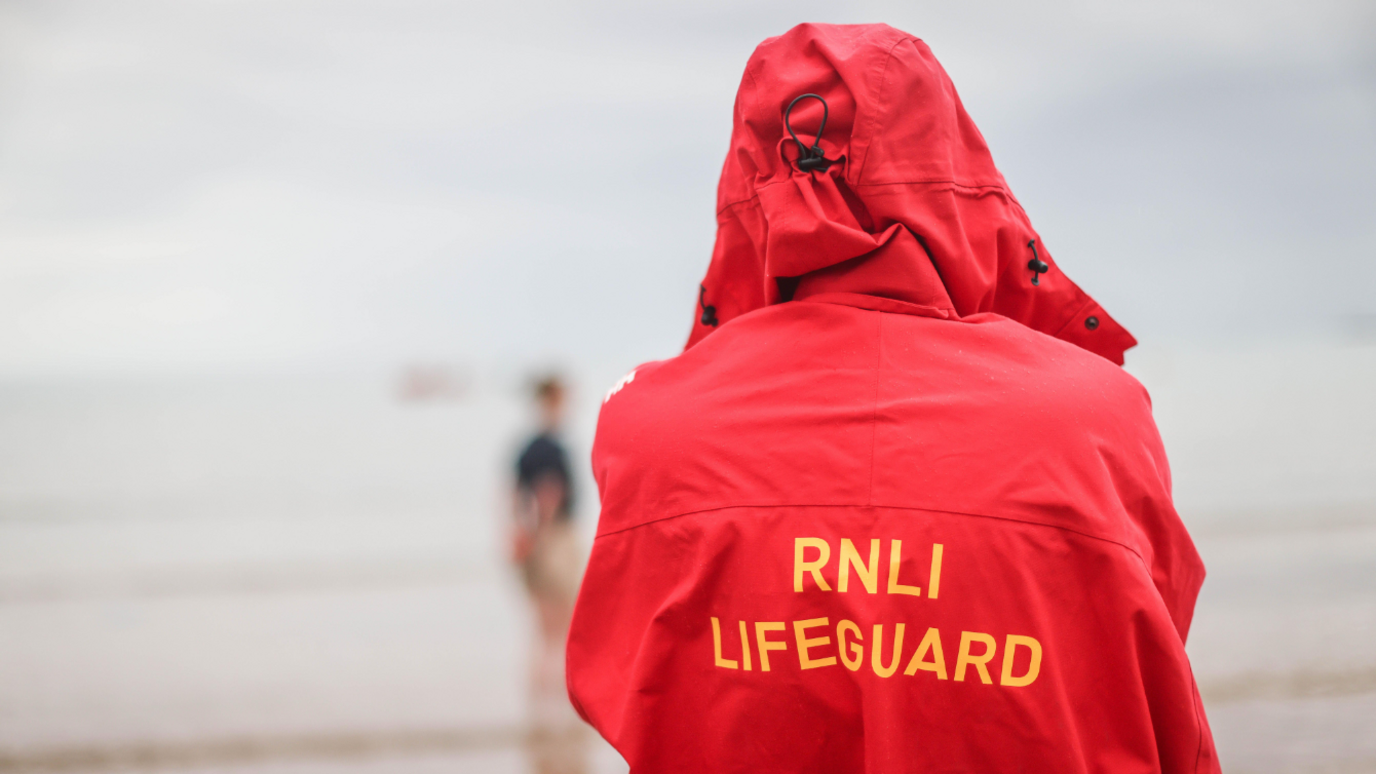 A person with their back facing the camera looking out to sea. They are wearing a red hooded coat which says RNLI Lifeguard in yellow capital letters