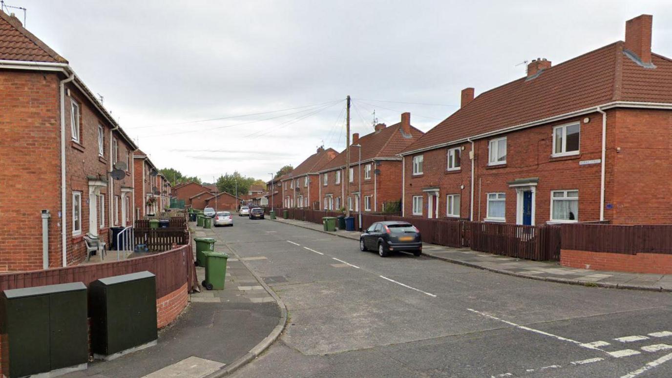 Morris Street in Gateshead. The picture is taken from the junction of the road and shows semi-detached housing lining the street. Several cars are parked outside and green wheelie bins have been put out for collection.