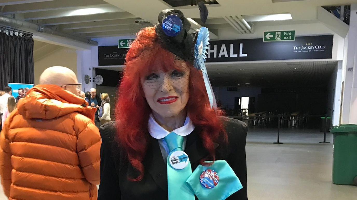 Gloria Jane Martin wears a black suit, a turquoise tie with a Reform UK badge pinned on and a black fascinator with another badge and Reform rosette attached,