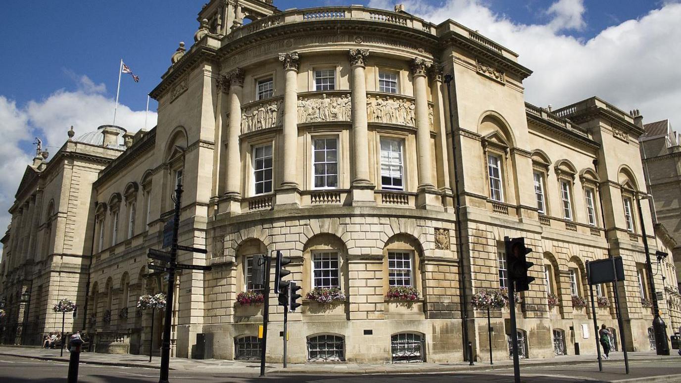 An historic old stone council building in front of a road under a blue, partially cloudy sky