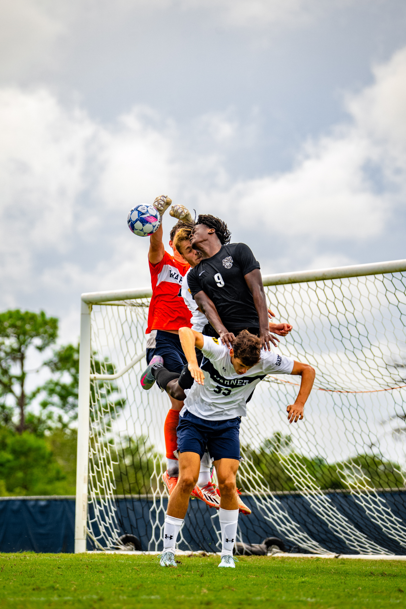 Four boys are trying to get the football in front of a goal. One player is using another's shoulders to jump to get to the ball with his head. There are two other players behind him also trying to jump for the ball.
